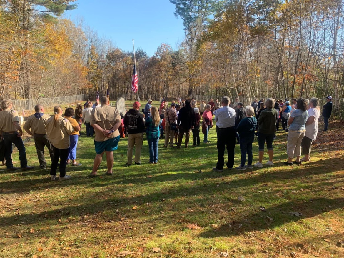 A large group of people gathered outdoors on a grassy field surrounded by trees, facing a flagpole with an American flag.