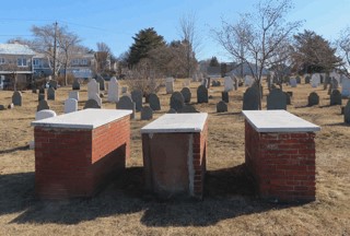 Three above-ground brick and stone tomb vaults with flat granite lids, set in a grassy cemetery with rows of upright headstones in the background