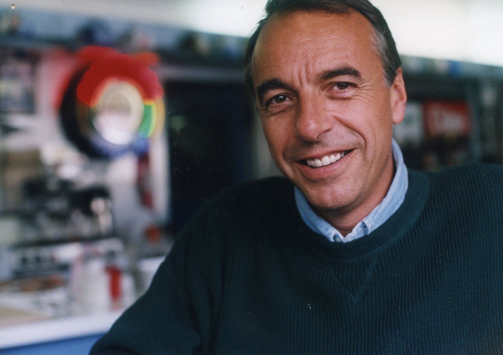 A man in a dark green sweater smiles at the camera while sitting indoors, with blurred shelves and colorful objects in the background.