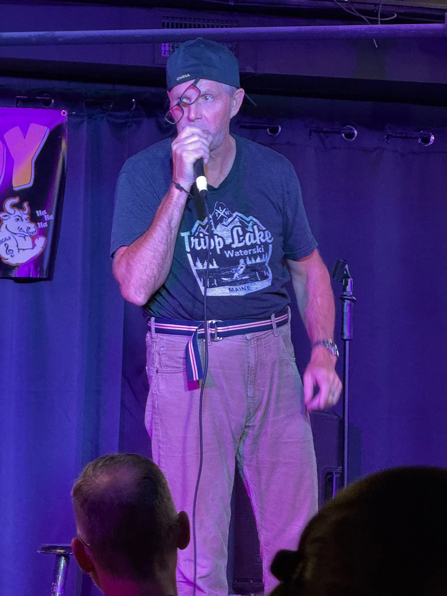 A man in a black shirt and hat tells jokes into a microphone on stage.