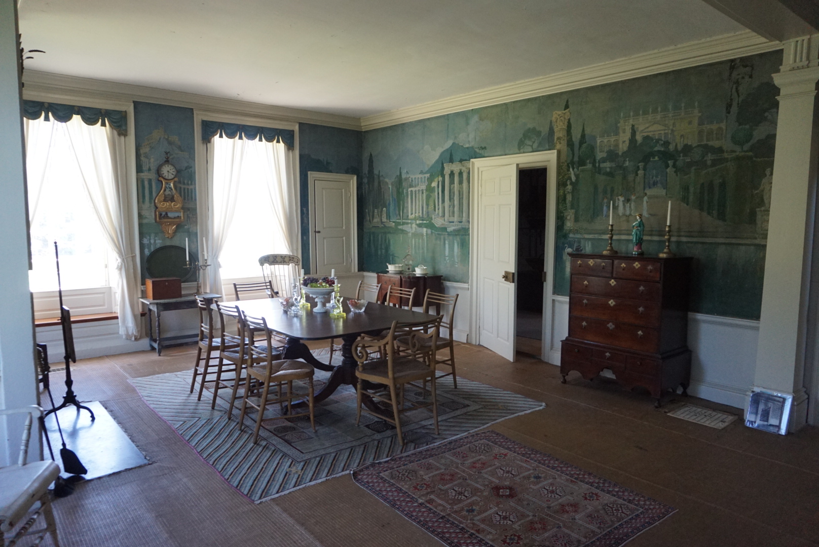 Historic dining room with a long wooden table and chairs, decorative hand-painted mural wallpaper depicting classical ruins, and an antique tall chest of drawers.