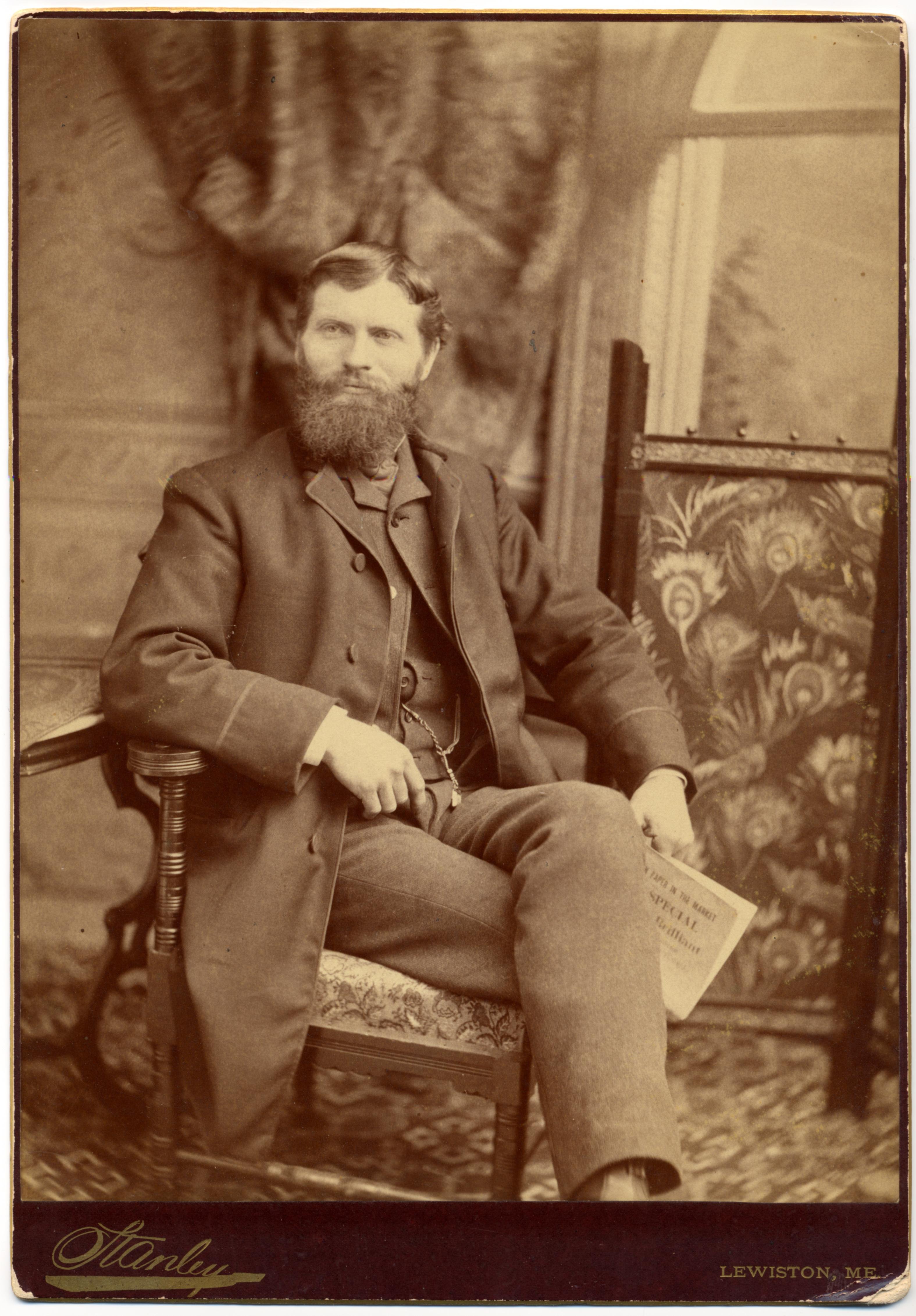 "Sepia cabinet-card portrait of a bearded man seated in an ornate chair, holding a folded newspaper, with a Stanley photography studio credit and 'Lewiston, ME' printed below, circa 1882."