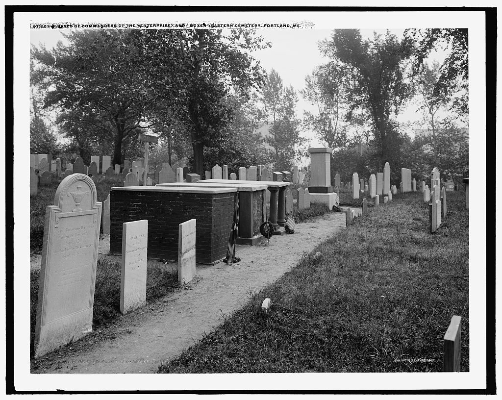 Black and white photo of a cemetery featuring various tombstones amidst overgrown grass and trees.