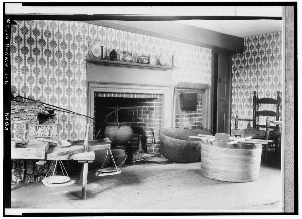 A historic black-and-white interior view of a kitchen featuring a large brick fireplace, patterned wallpaper, wooden tubs, and period cookware.