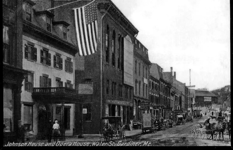 Black-and-white historic postcard photo of "Johnson House and Opera House" on Water Street in Gardiner, Maine, showing horse-drawn carriages on the street and a large American flag hanging from the building.