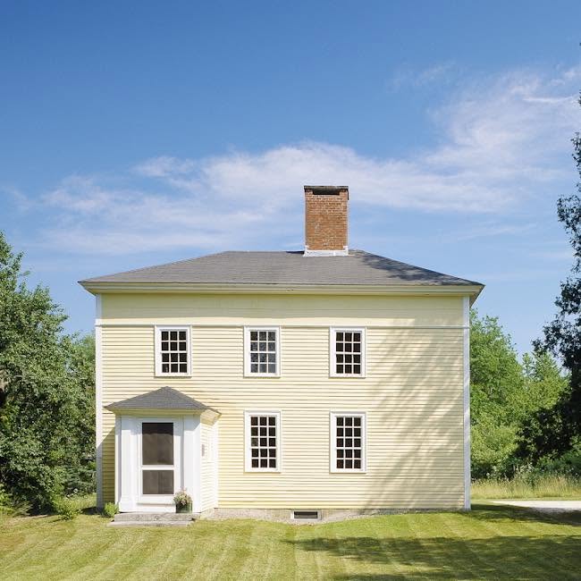 A yellow colonial-style farmhouse with a central brick chimney and gray shingle roof.