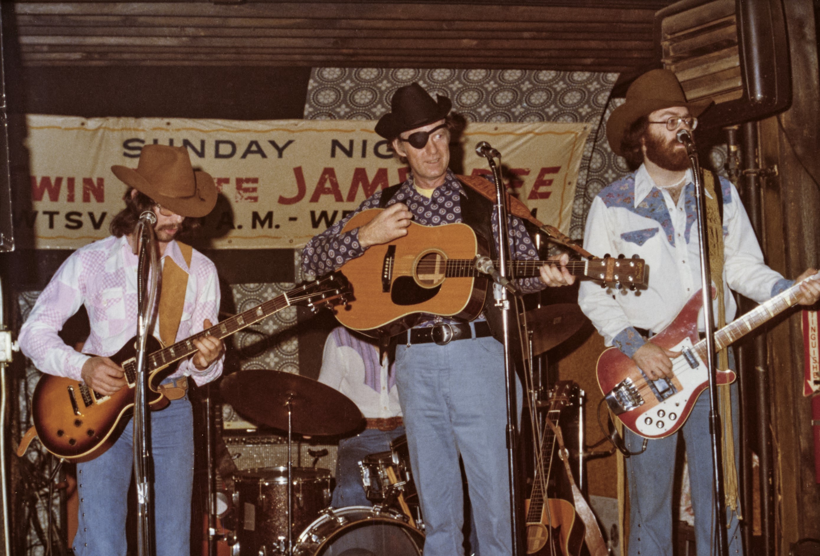 Vintage color photo of the same band performing live, singer in cowboy hat and eye patch at center mic with acoustic guitar, flanked by guitarist and bassist; banner reads "Sunday Night Jamboree, WTSV AM-FM."
