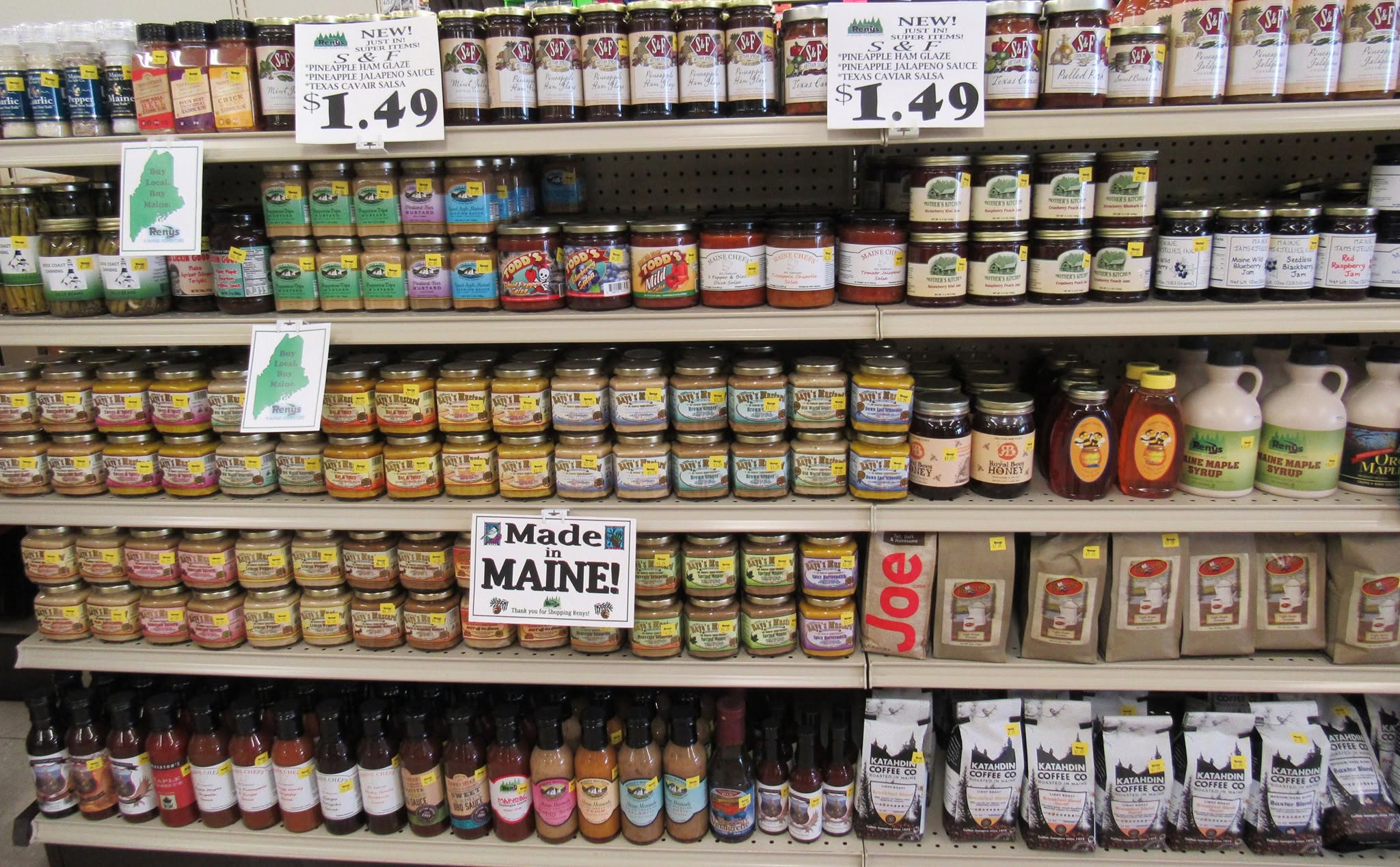Grocery store shelves stocked with Maine-made products including mustards, jams, hot sauces, honey, maple syrup, and coffee, with signs reading "Made in Maine!" and "Buy Local, Buy Maine."