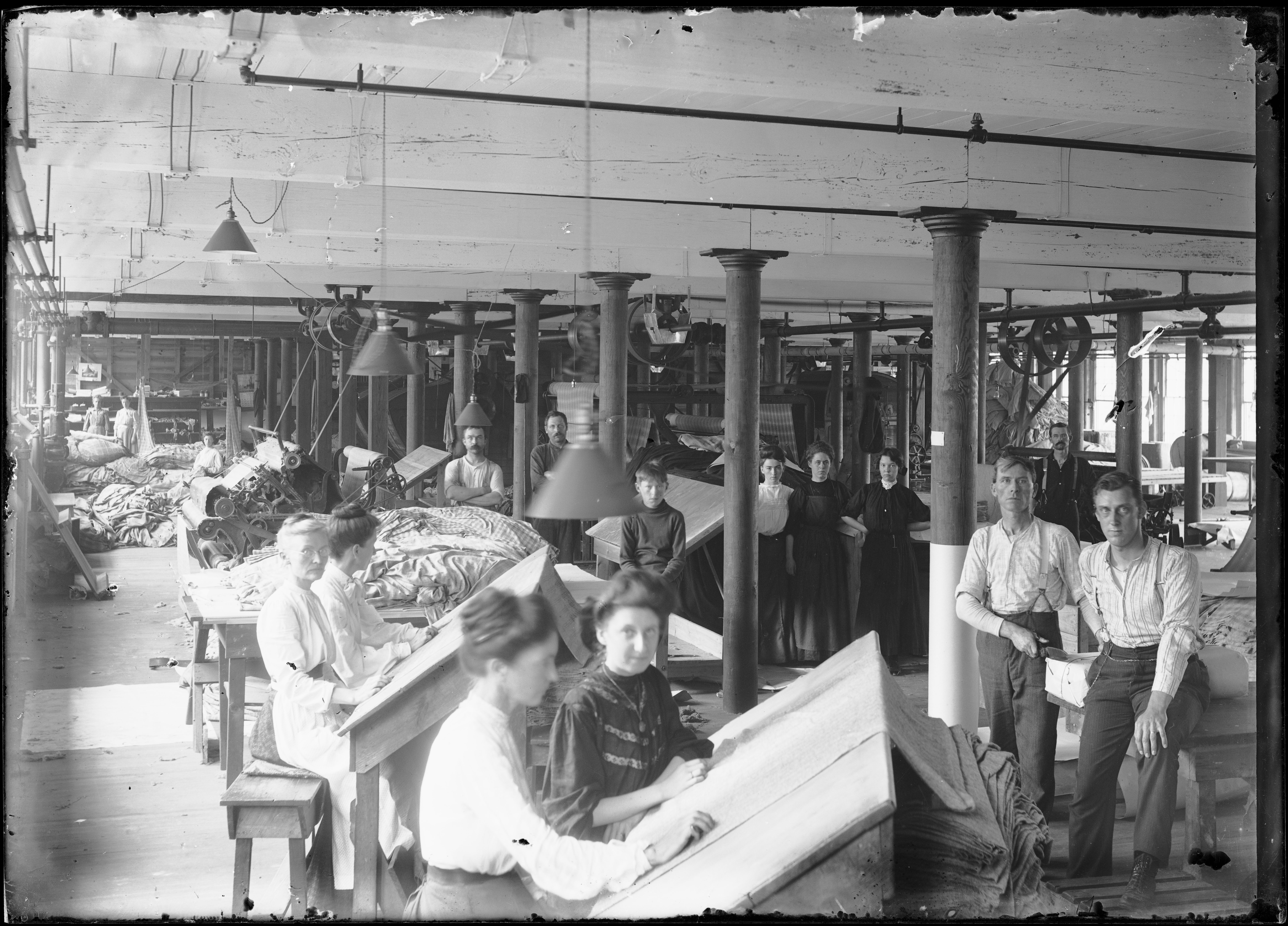 Vintage black and white photo of textile mill workers, including women inspecting cloth at slanted tables and men standing among machinery, columns, and stacked fabric rolls in an early 20th-century factory interior.