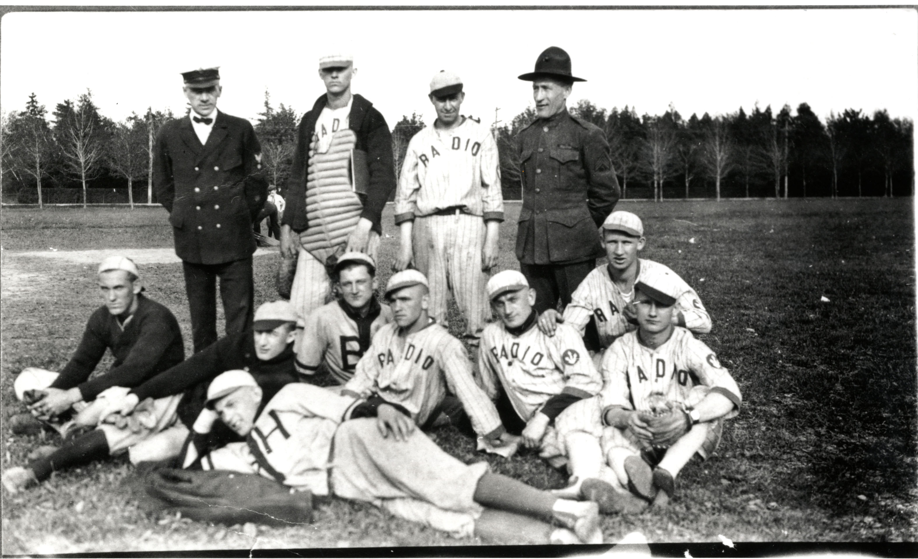 Historic black-and-white photo of the Otter Cliffs Radio Station baseball team, players in "Radio" jerseys posed sitting and standing on a grass field with a treeline in the background.