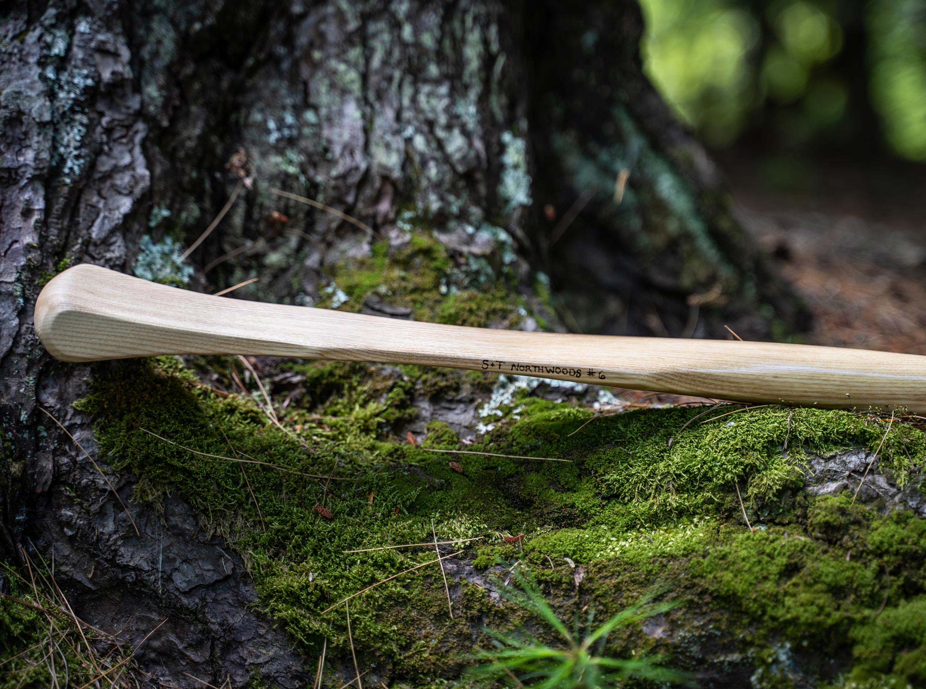 Close-up of a wooden canoe paddle blade stamped "S+T Northwoods #6," resting on a moss-covered log against a tree trunk in the woods.