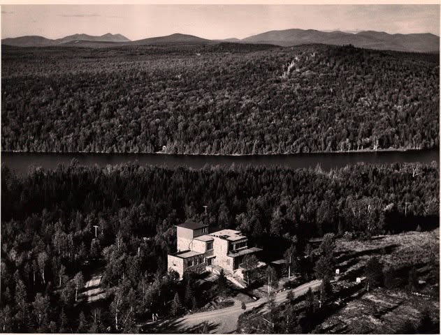 "Aerial black-and-white photo of a stone building on a hillside overlooking a lake, surrounded by dense forest with mountains in the distance."