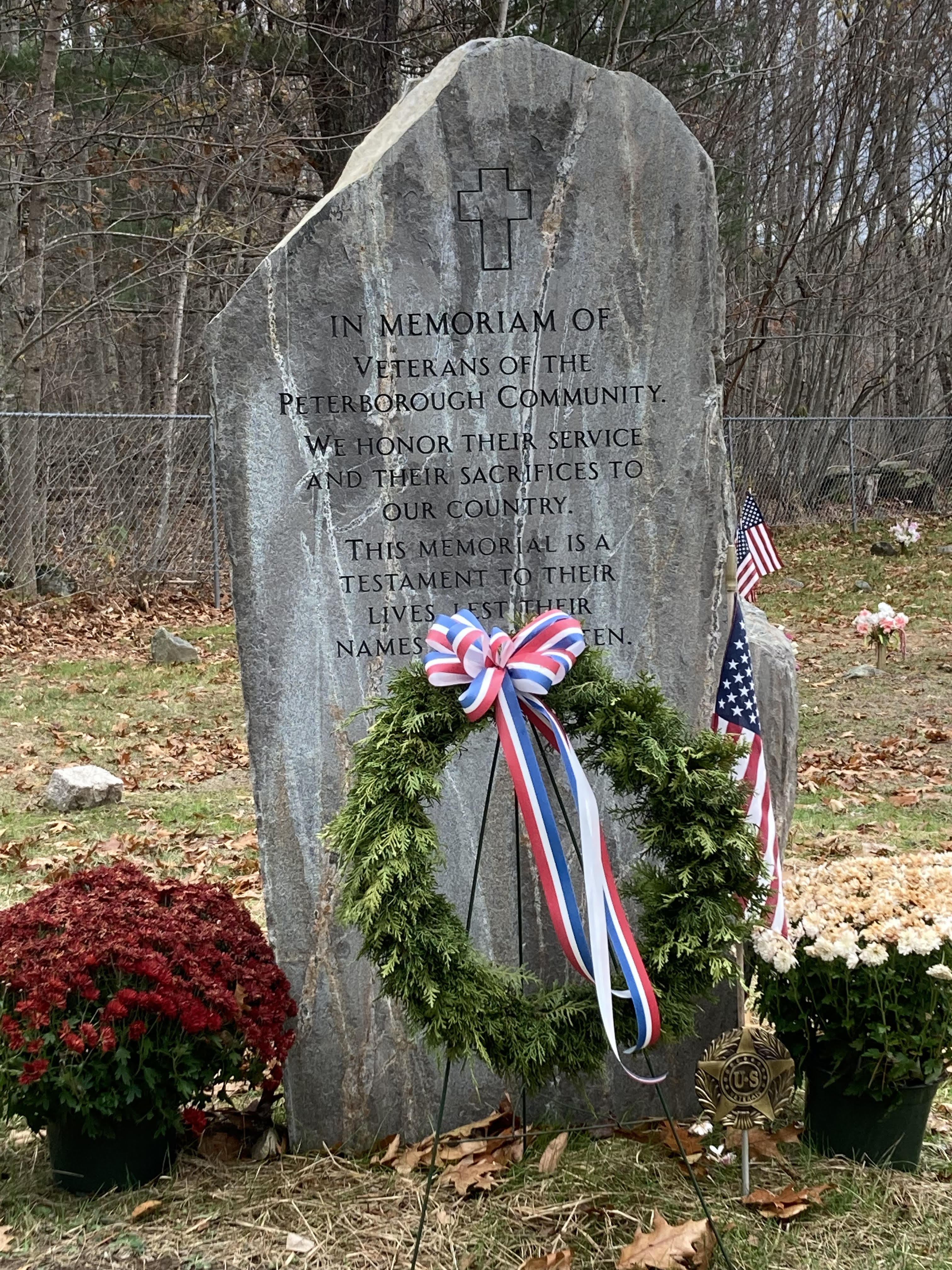 The Peterborough Veterans Memorial stone decorated with a evergreen wreath featuring a red, white, and blue bow, flanked by potted mums and American flags.