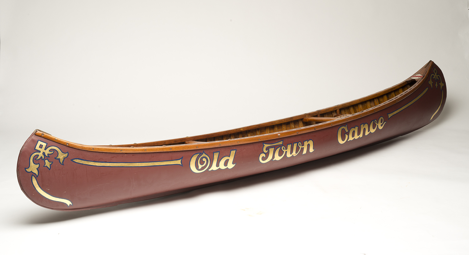Side view of an antique wooden canoe with dark red hull and ornate gold scrollwork lettering reading "Old Town Canoe," photographed against a plain white background.