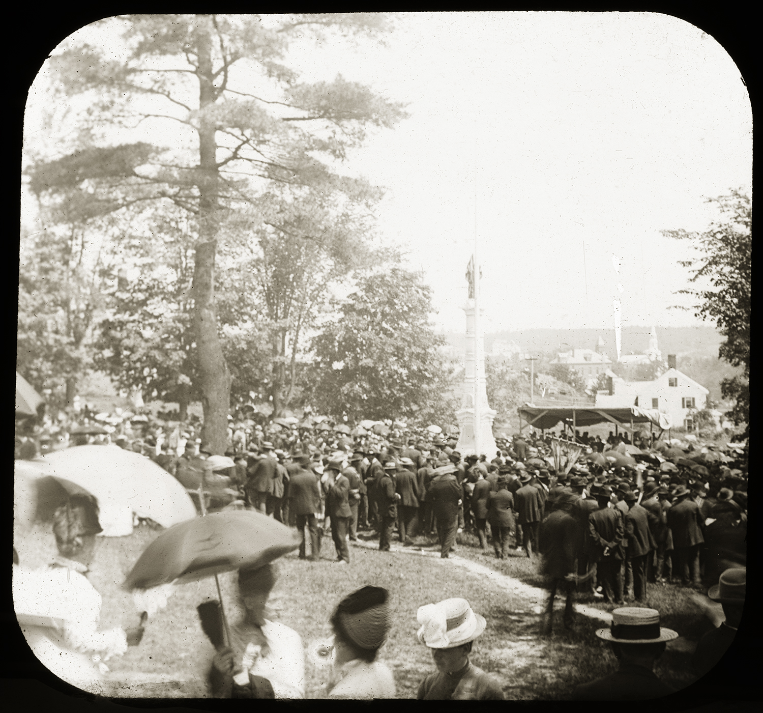 Historic photo of a large outdoor crowd gathered around a tall monument on a hillside, with women in the foreground holding parasols and a town visible in the background.