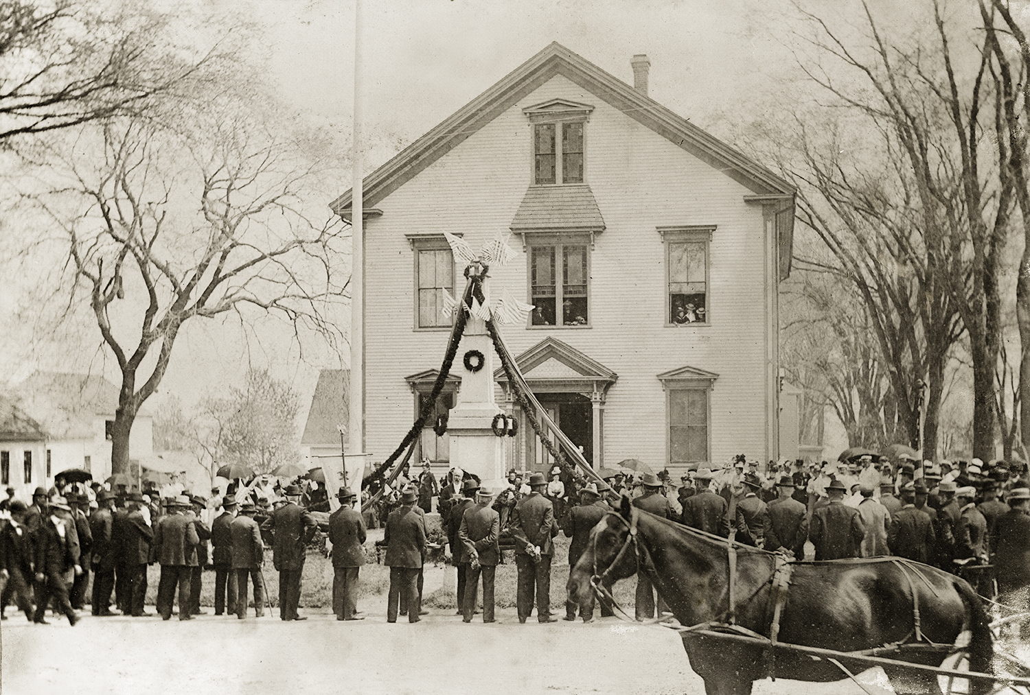 Historic sepia-toned photo of a crowd of people and a horse-drawn carriage gathered in front of a large white house for a monument dedication, with a flag-draped memorial and wreaths visible near the entrance.