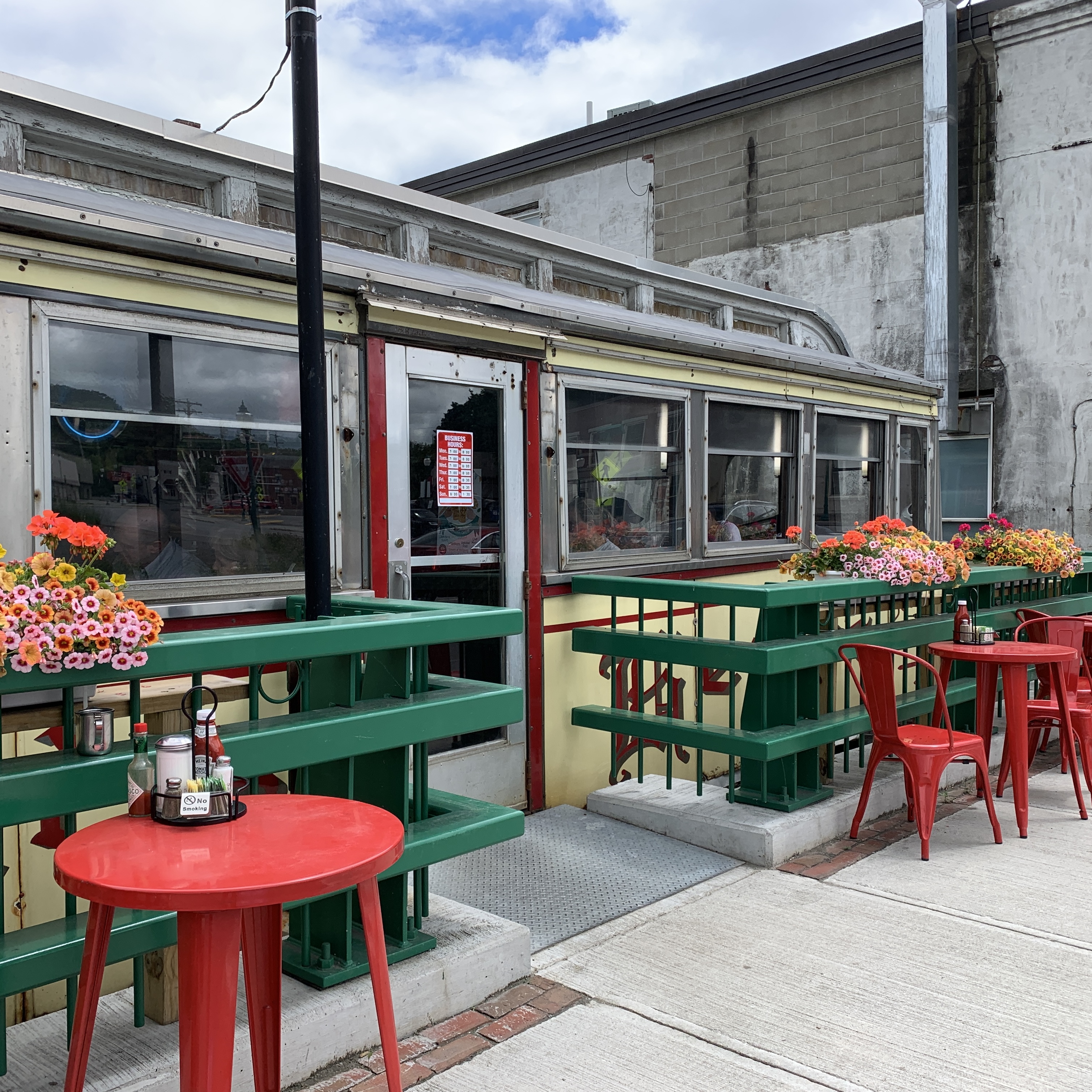 Exterior of a vintage diner car with red trim, viewed from an outdoor patio with green railings, red bistro tables and chairs, and hanging flower boxes.
