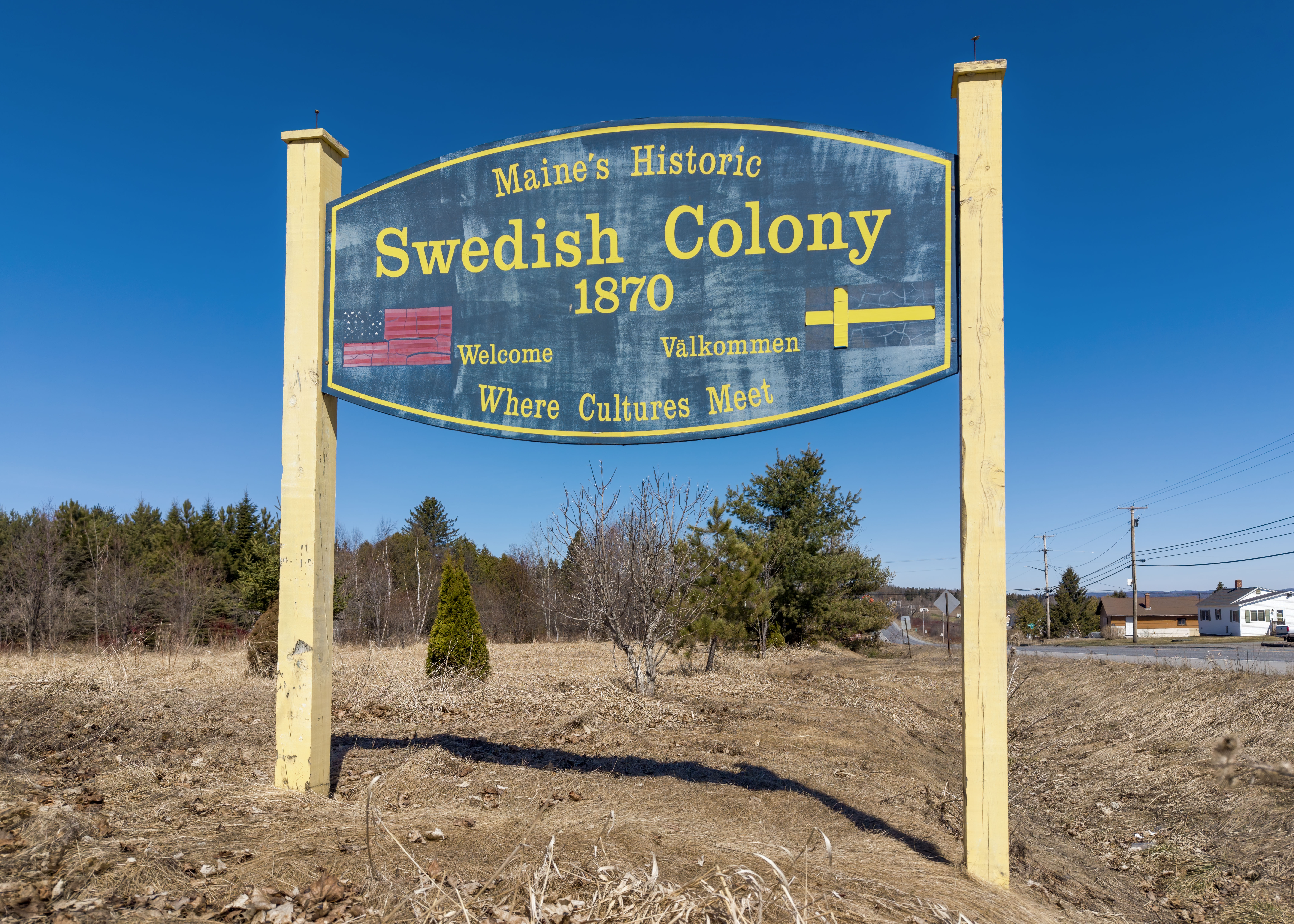 An outdoor sign reading "Maine's Historic Swedish Colony 1870" flanked by two yellow posts, set against a blue sky and a dry field.