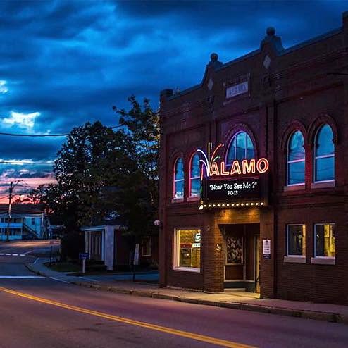 Twilight photo of the brick Alamo Theatre with a glowing neon marquee sign reading "Alamo," arched windows, and a movie listing on the marquee, situated on a small-town street corner.