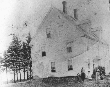 A vintage black-and-white photograph of several people standing in front of a large multi-story historic house near a row of tall evergreen trees.