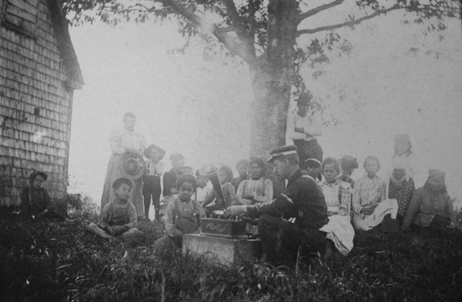 A historical black-and-white photograph of Jesse Walter Fewkes operating an early phonograph outdoors for a gathering of people seated on the grass near a building and a tree.