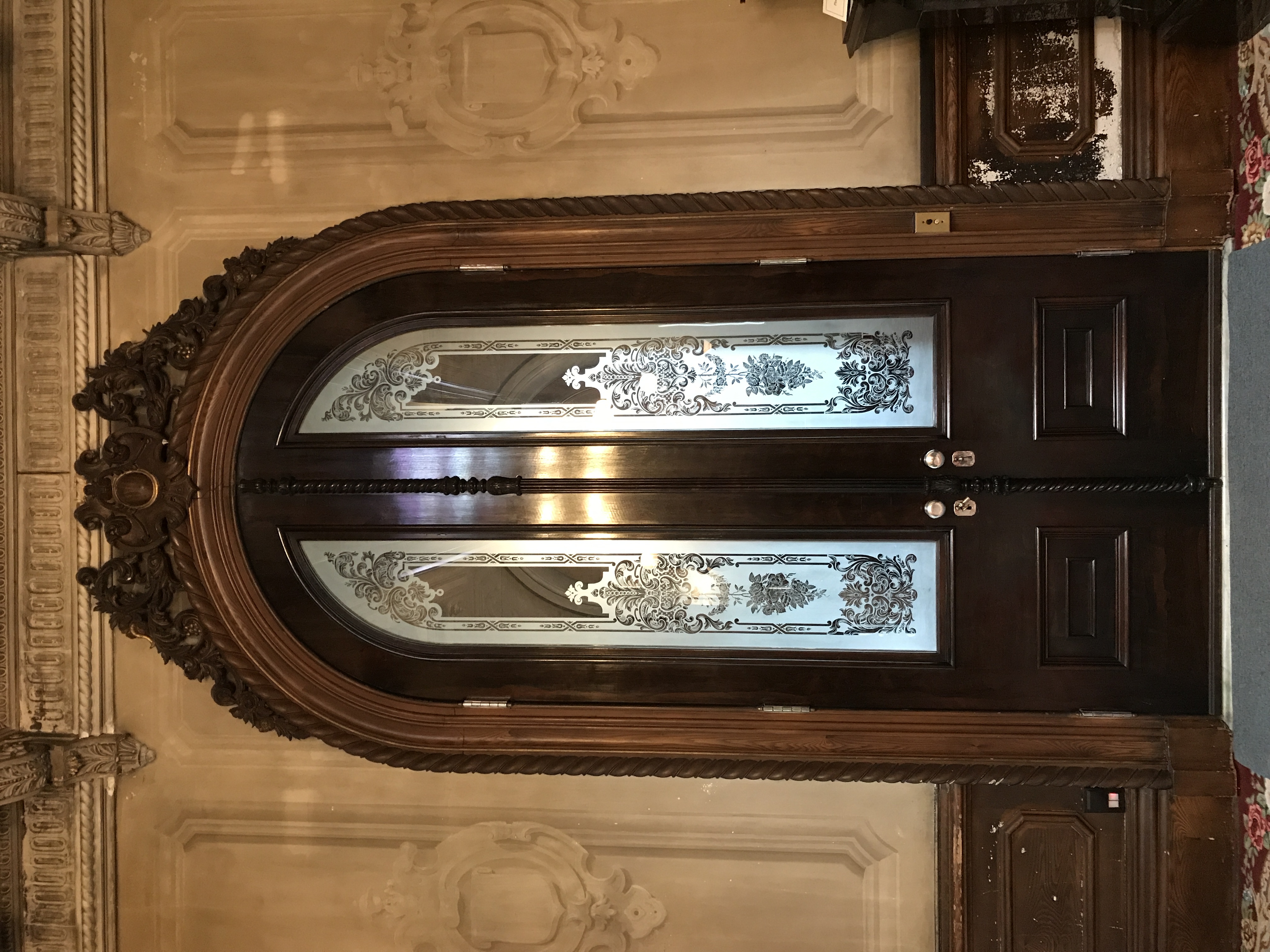 A pair of tall, dark wooden double doors with arched tops and intricate frosted glass panels, set into a wall with carved molding and neutral-toned paneling.