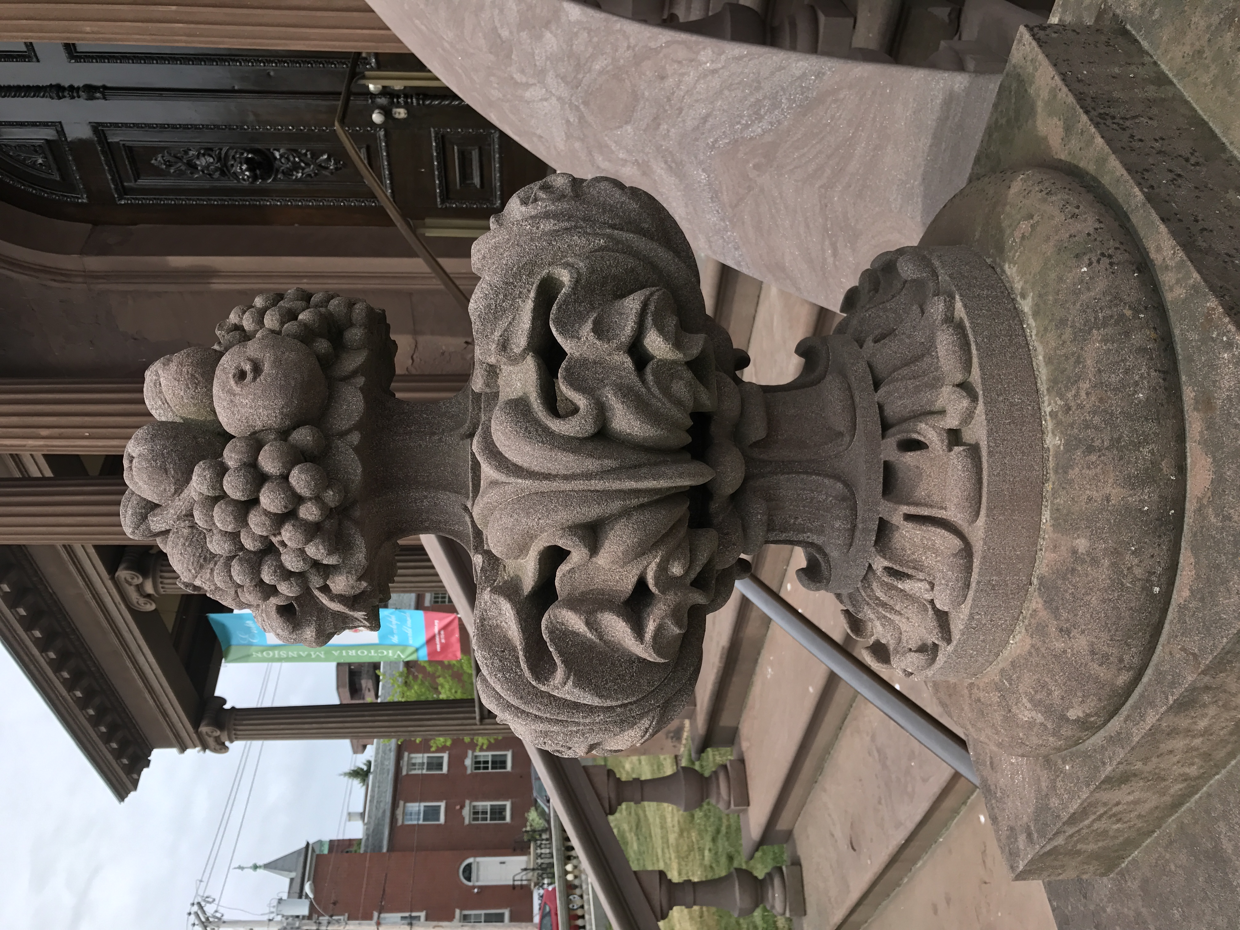 A close-up shot of an ornate stone architectural sculpture shaped like a carved urn filled with fruit and leaves, sitting on a pedestal outside a building.
