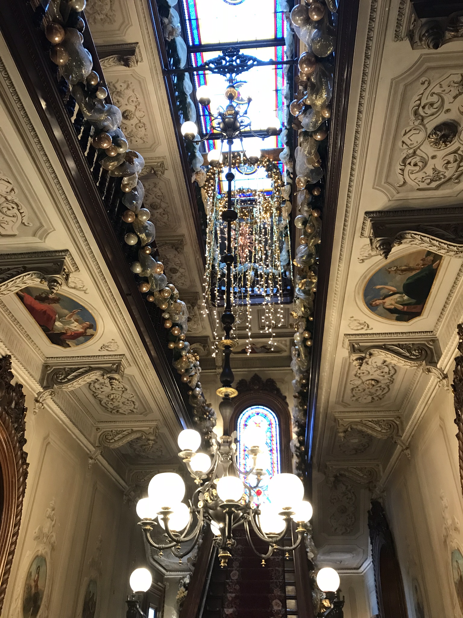 A vertical view looking up a decorated staircase with holiday garland along the banister, a multi-tiered chandelier, and framed ceiling paintings.