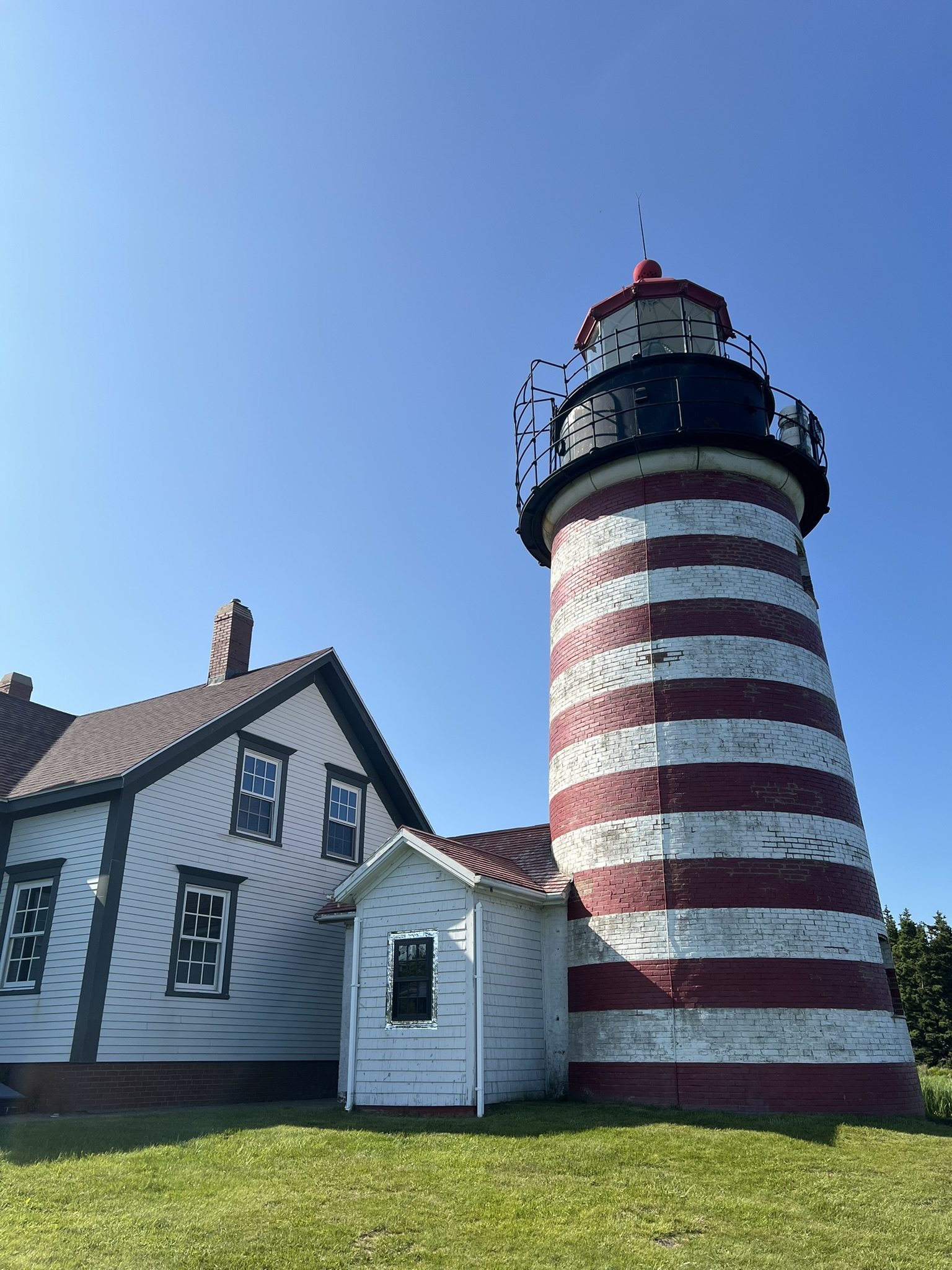 A low-angle view of a red-and-white horizontally striped lighthouse next to a white house with a dark roof against a clear blue sky.