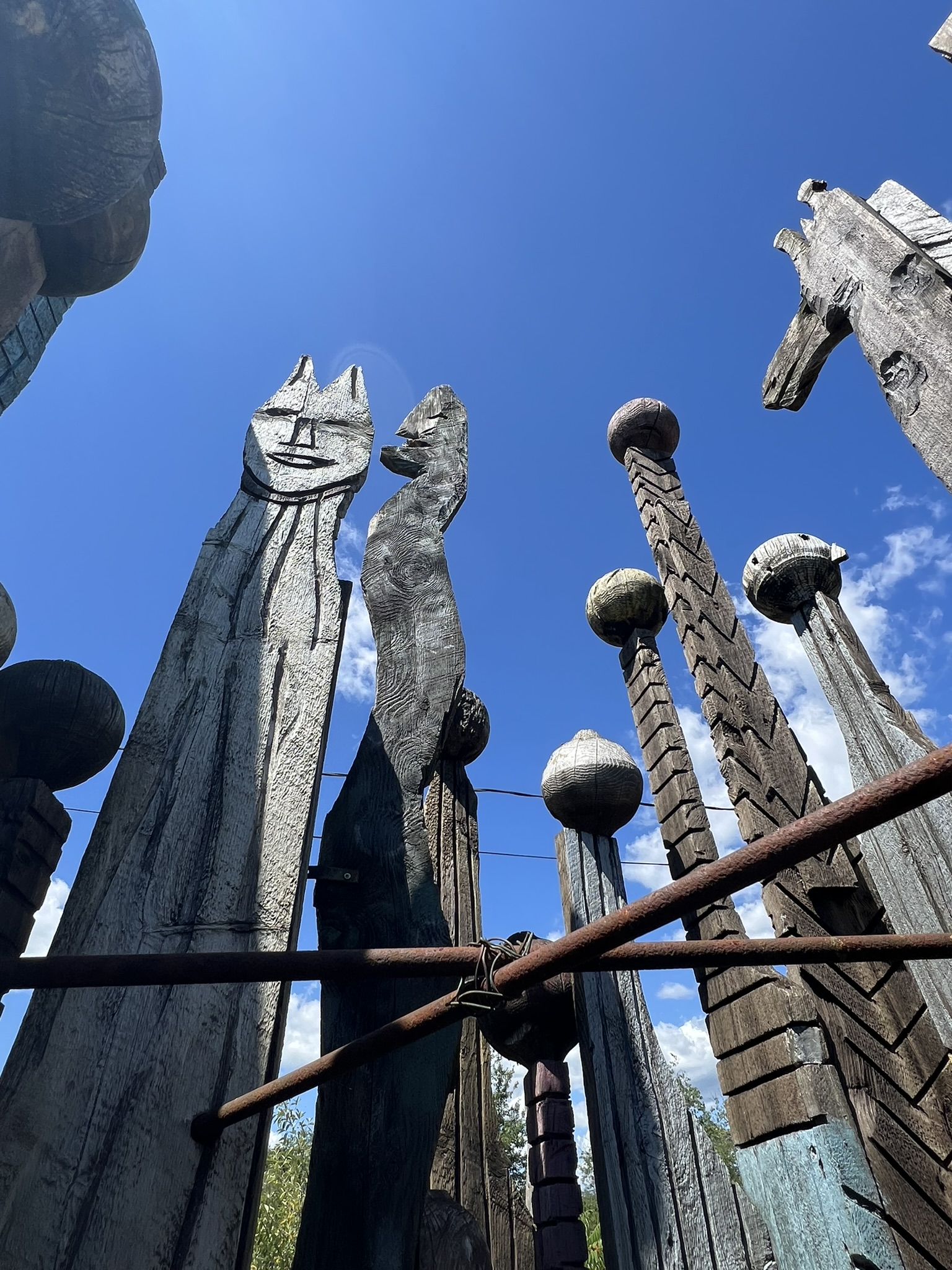 Low-angle view of tall, weathered wooden sculptures with carved faces and spherical finials against a bright blue sky, with a horse figure visible at right.