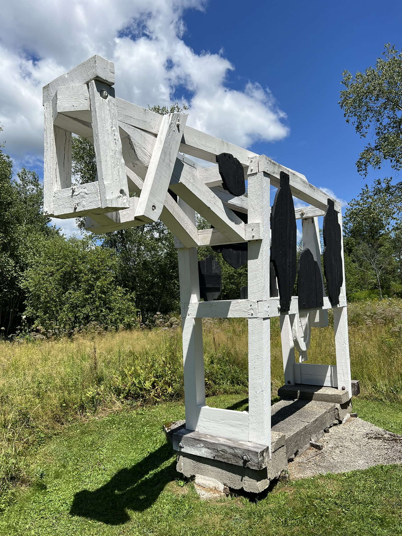 White-painted wooden sculpture resembling an abstract horse or animal frame with black paddle-like shapes, standing in a grassy field.