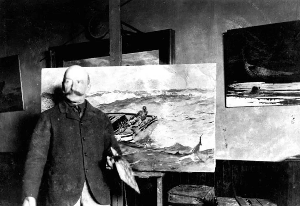 Black-and-white historical photo of artist Winslow Homer standing in his studio holding a palette, with one of his large seascape paintings on an easel behind him.