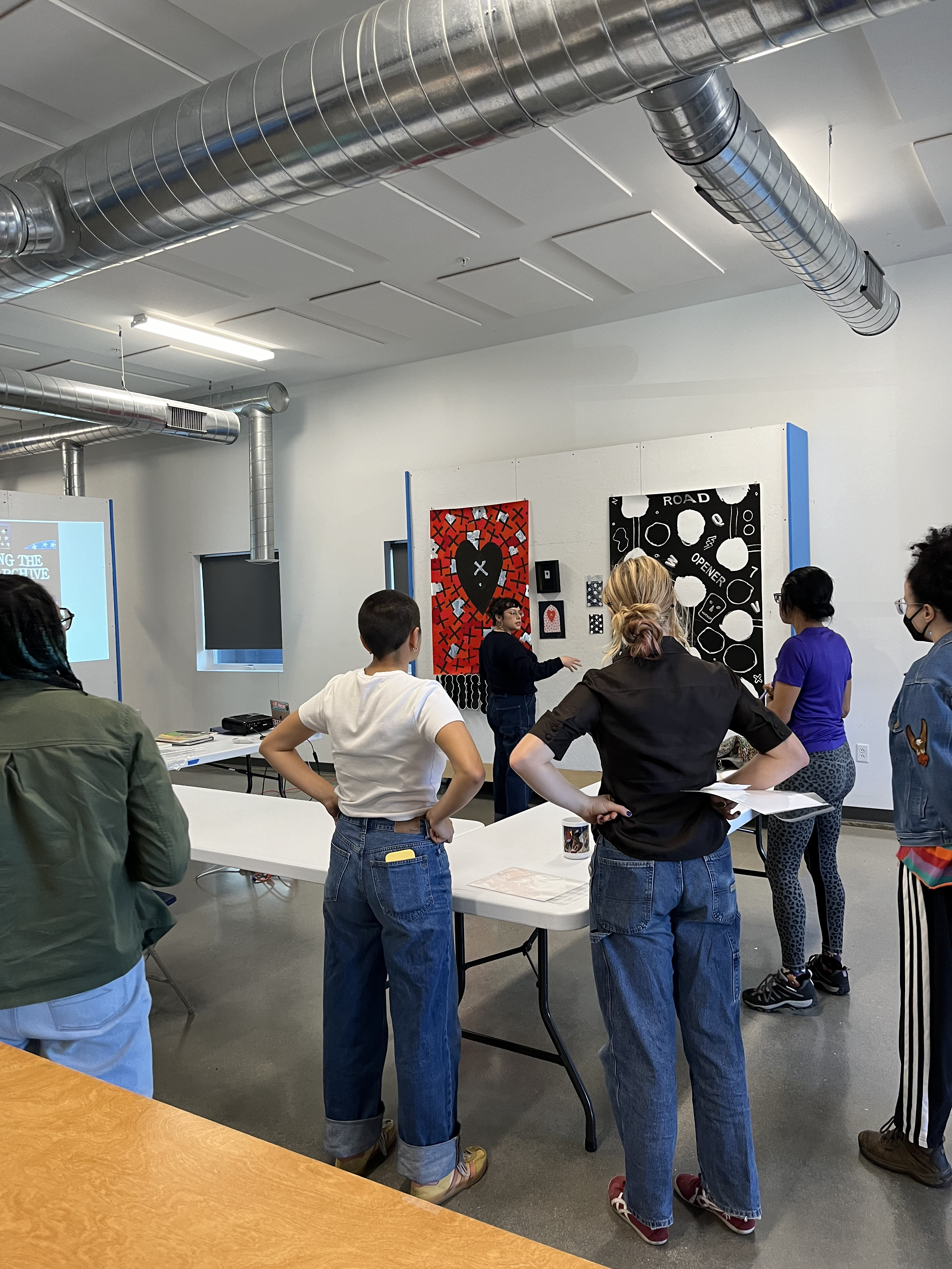 A group of people stand in a bright, modern room facing a wall displaying framed art, listening to an instructor.