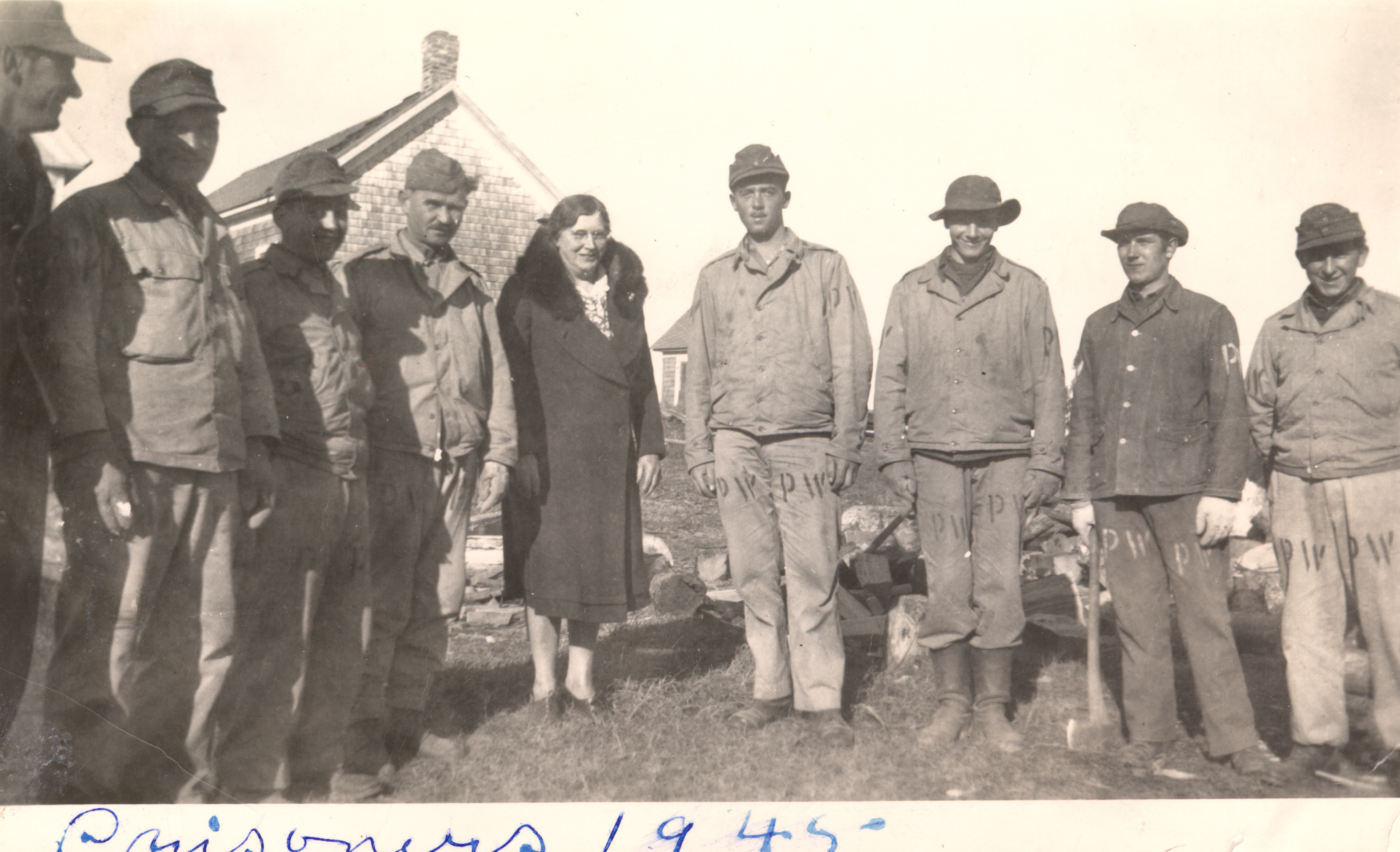 A vintage black-and-white photo from 1945 showing a woman standing outdoors with a group of men, several wearing uniforms with "PW" stenciled on their trousers.
