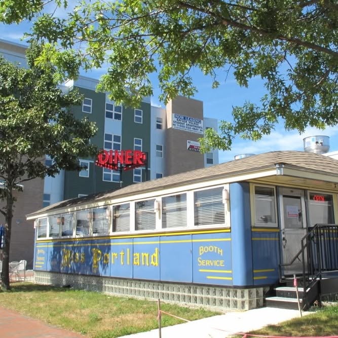 Exterior of a vintage diner car with red trim, viewed from an outdoor patio with green railings, red bistro tables and chairs, and hanging flower boxes.