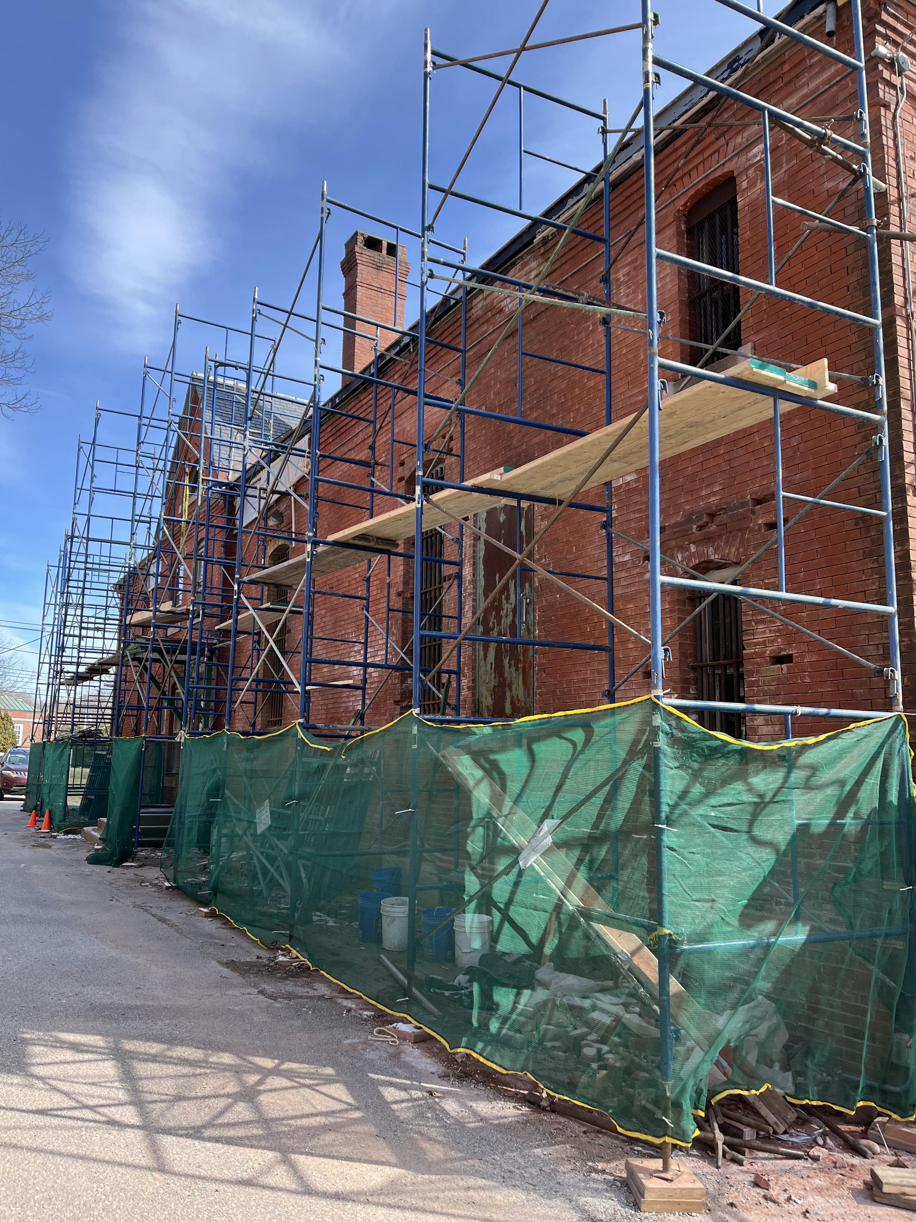 A multi-story blue scaffolding structure erected alongside the exterior wall of a red brick building, partially covered with green protective mesh at the base.