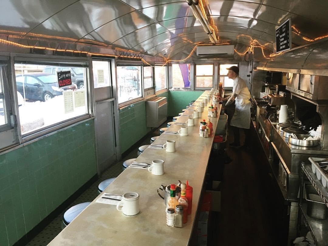 Interior of a narrow diner car with a long counter, blue coffee mugs, condiment bottles, string lights along the curved ceiling, and a cook working at the grill.