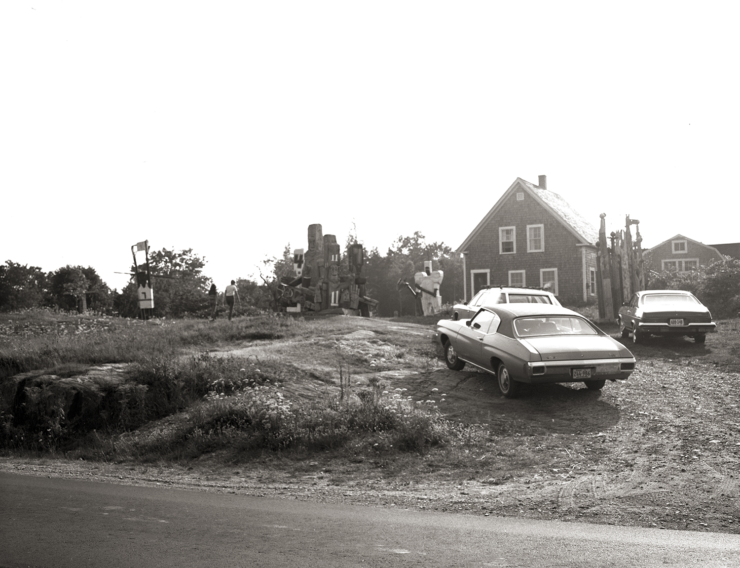 Black-and-white photo of a roadside sculpture garden with tall carved wooden figures near a shingled house, with two vintage cars parked on the gravel in front.