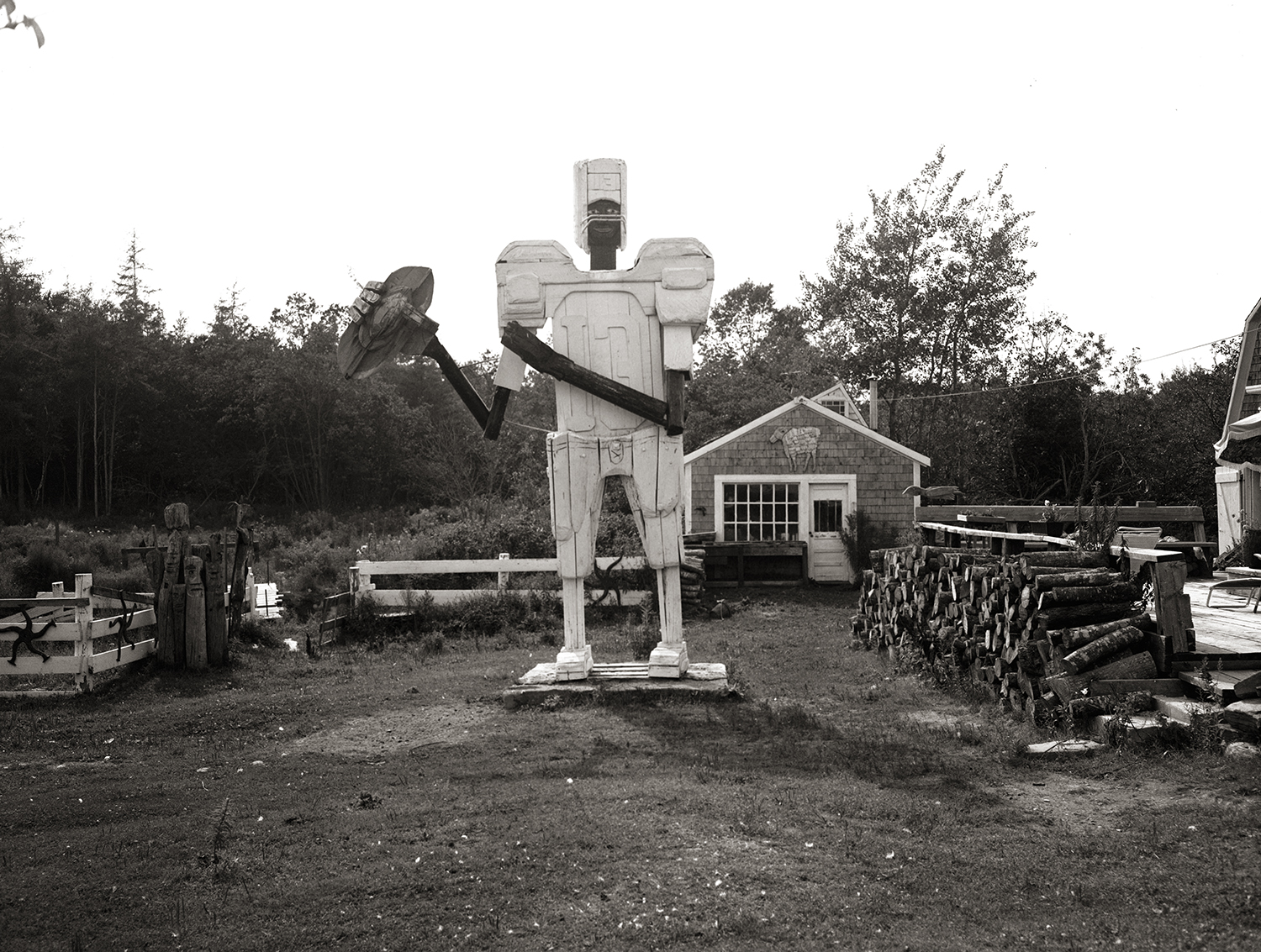 Black-and-white photo of a tall wooden robot-like sculpture holding an axe, standing in a yard near a shingled shed, fencing, and a stacked woodpile.