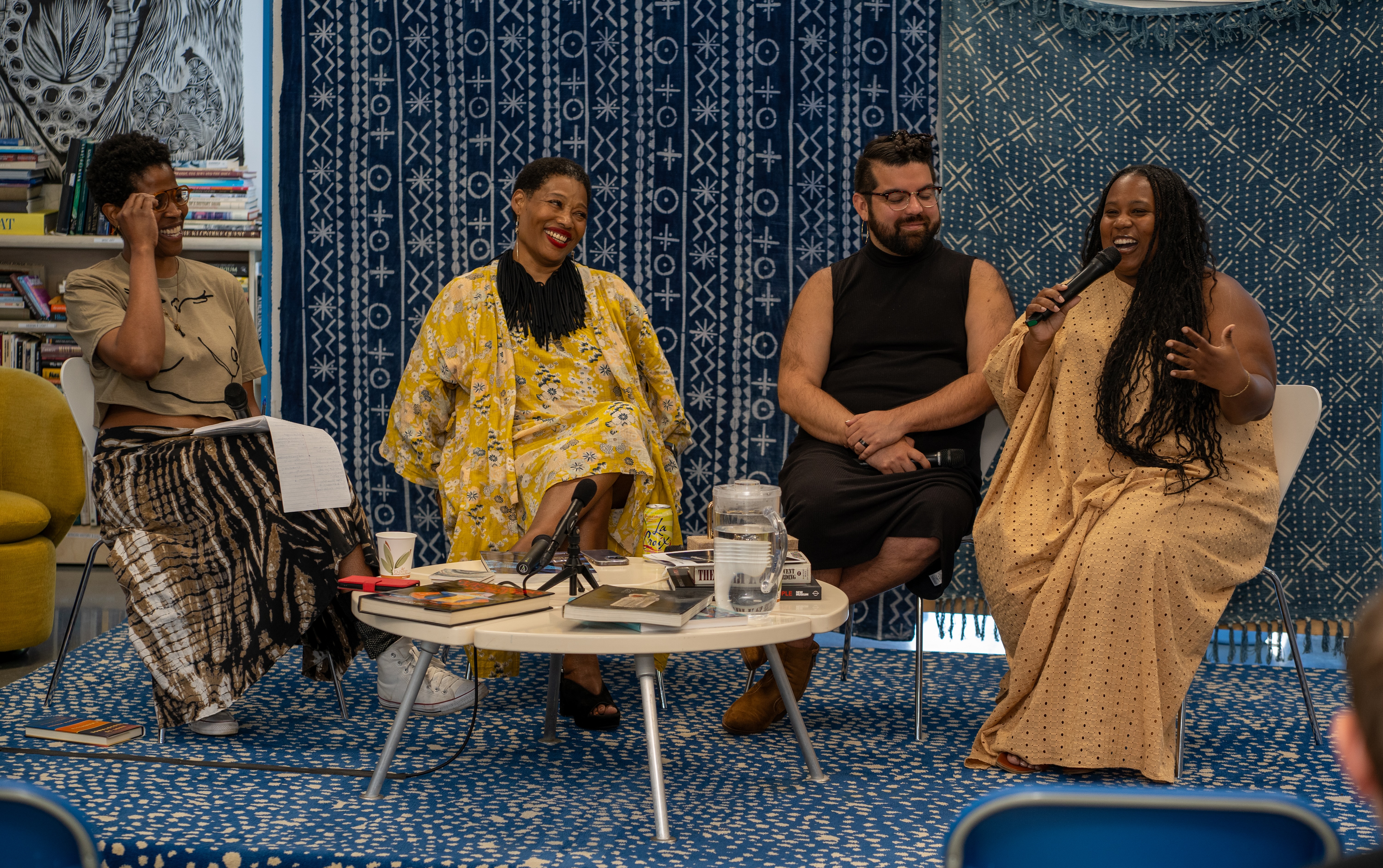 Four panelists seated around a table on a blue-and-white patterned rug, speaking into a microphone in front of a patterned indigo backdrop.
