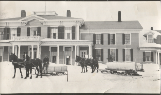 Historic black-and-white photograph of the large wooden estate in winter, featuring horses harnessed to sleds or carriages on the snow-covered grounds.
