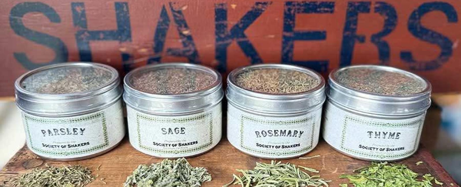 Four glass jars of clear herbs with wooden plank labeled Shakers in background