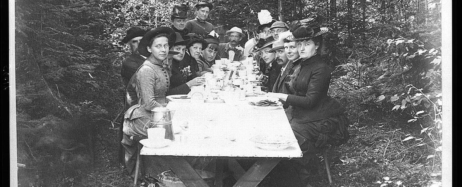 A black and white historical photograph depicting a large group of men and women dressed in Victorian-era attire. They are seated along a long wooden picnic table covered in a white tablecloth, dining inside a dense forest clearing. Handwritten names are visible on the top and bottom margins of the photo.