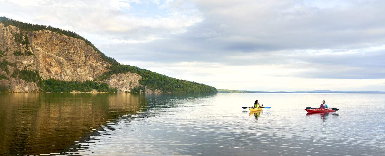 A scenic landscape photograph showing two kayakers—one in a yellow kayak and one in a red kayak—paddling on calm, wide water. On the left, a massive, sheer rocky cliff face rises up from the shoreline under a cloudy blue sky.