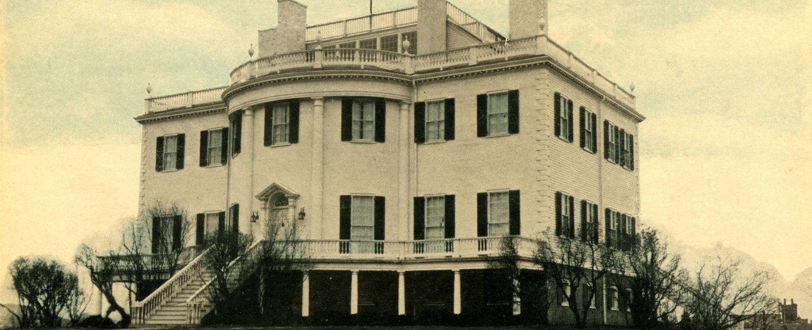 A vintage, lightly tinted historical photograph of the white Montpelier estate viewed from a three-quarters angle. The three-story mansion features a large curved front section, extensive balustrades, and dark shutters, surrounded by a grassy lawn and bare trees.