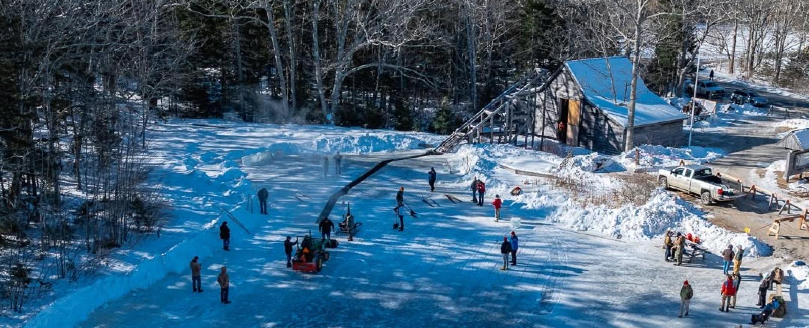 Aerial view of a snow-covered pond where a crowd harvests ice in long cut rows, near a weathered barn with a blue roof and parked truck.