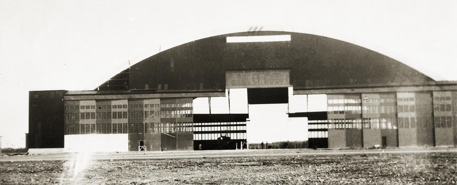 Vintage black and white photo of an arched airplane hangar at Limestone Air Base, Maine, with rows of windows along its base and open dirt field in the foreground, captioned "Hanger, Limestone Air Base, Limestone, ME."